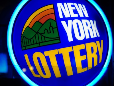 A bright, circular neon sign with a blue background, featuring the words 'New York Lottery' in bold white and yellow letters. The sign includes a colorful illustration of a landscape with green mountains, an orange-red sunset, and a bridge, all encapsulated within the circle.