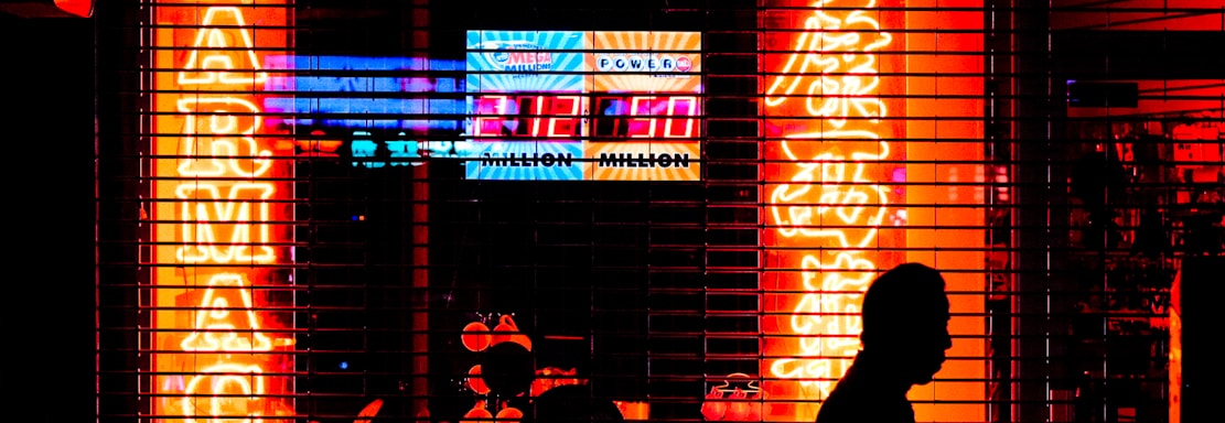 Bright neon signs in orange and yellow light up the storefront of a pharmacy, visible through metal security bars. The silhouette of a person walking by is prominent in the foreground, set against the vibrant colors. Additional signage displays a lottery number in bright white and blue.
