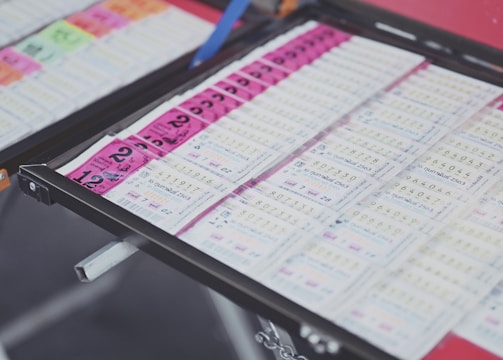 A collection of lottery tickets is organized neatly in a metal tray. The tickets display various numbers and have color-coded sections, primarily in pink and white. Some tickets have vibrant green sections, adding contrast to the overall layout.