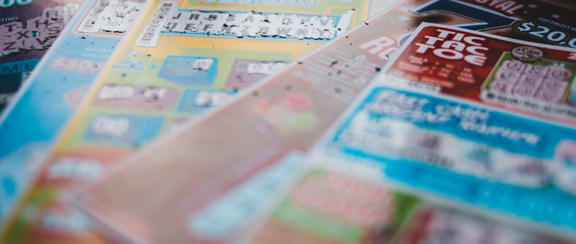 A selection of colorful lottery scratch-off tickets placed on a flat surface near a window. The image has a shallow depth of field, focusing on the tickets while blurring the background. The tickets display various games and prize amounts.