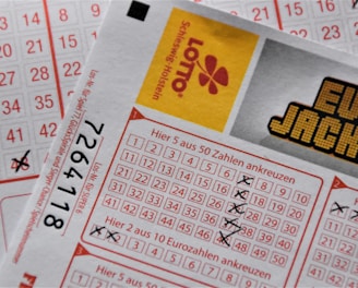 A close-up image of lottery tickets featuring crossed-out numbers, brand logos, and various numbers in a grid format. The tickets have sections for different games, and numbers are marked with black crosses.