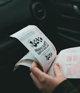 A person is holding and examining what appears to be lottery tickets inside a car. The image is focused on the tickets and the person's hands, with parts of the car interior visible in the background.