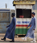 Two individuals are walking past a small kiosk with signage for the National Lottery Authority. The kiosk, decorated with a sign that reads 'Full Payment,' features yellow, green, red, and white colors. The individuals are dressed in matching long blue robes and hats, with one wearing black boots and the other white boots.