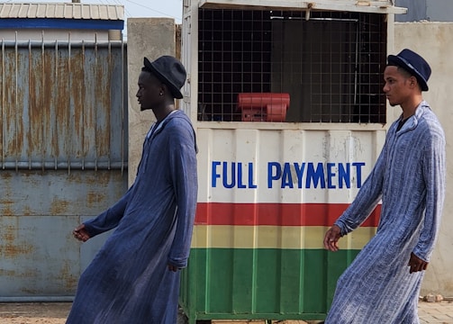 Two individuals are walking past a small kiosk with signage for the National Lottery Authority. The kiosk, decorated with a sign that reads 'Full Payment,' features yellow, green, red, and white colors. The individuals are dressed in matching long blue robes and hats, with one wearing black boots and the other white boots.