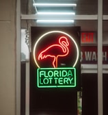 A neon sign depicting a flamingo above the words 'Florida Lottery' is prominently displayed. The sign is illuminated in bright red and green colors, and it is framed by glass panels. An 'EXIT' sign is visible in the background.
