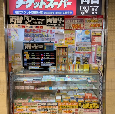 A storefront with various promotional banners and signs in multiple languages advertising ticket exchange services. The counter displays numerous tickets and small items, while the walls are covered with colorful posters and price listings.