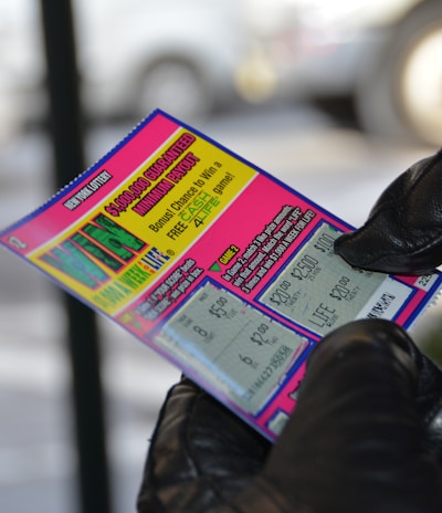 A person wearing black gloves is holding a colorful lottery scratch-off ticket. The ticket includes information about a $1,000,000 guaranteed minimum payout and features multiple games with various prize amounts.